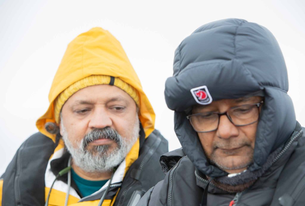 Two men in warm, layered jackets and hats, standing close together in a cold, outdoor environment.
